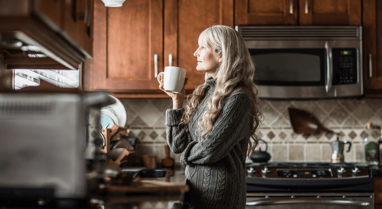 An older woman is standing in her kitchen with a mug, she is staring out a window that is mostly off of the screen